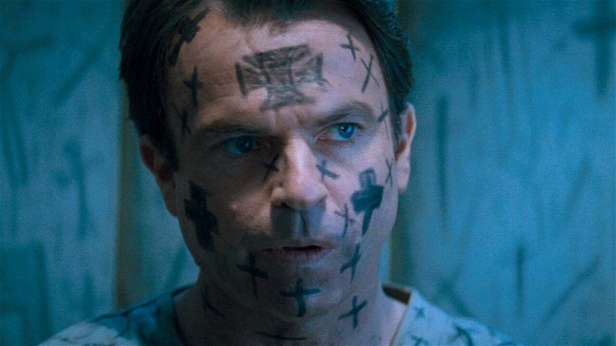 John Trent (Sam Neill) con la faccia ricoperta di croci in un manicomio