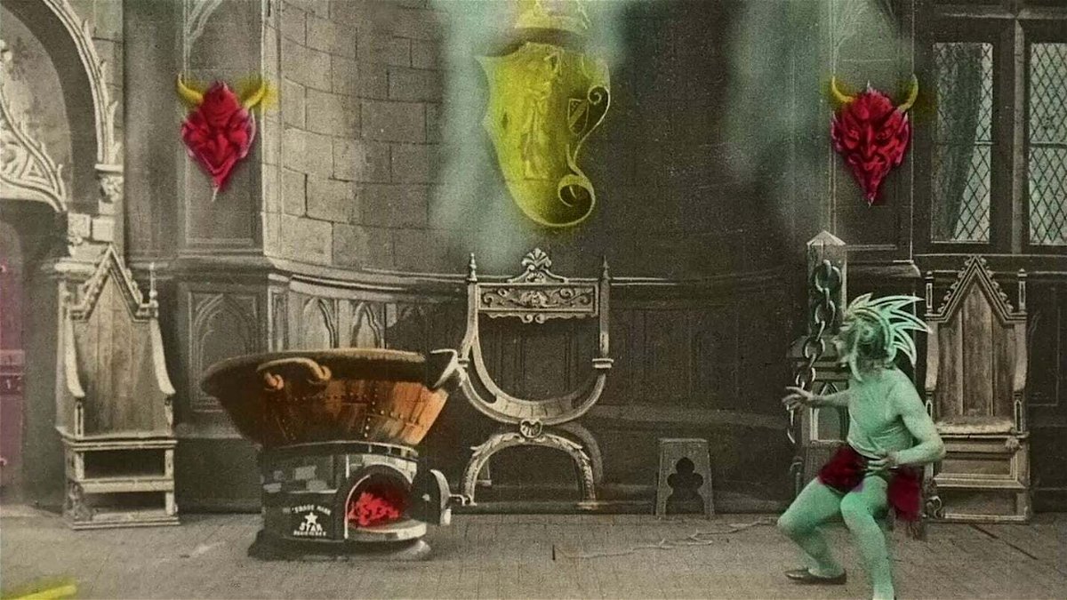 Un satanasso guarda un calderone che bolle in una sala antica. La pellicola in bianco e nero è colorata a mano.